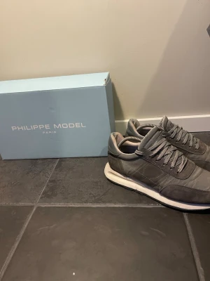 Grå Philippe Model sneakers - Snygga grå sneakers från Philippe Model med ovandel i mocka och textil, vita sulor och klassisk låg profil. Skorna har matchande grå skosnören och diskreta sömdetaljer. Perfekta för dig som gillar stilrena och exklusiva sneakers.