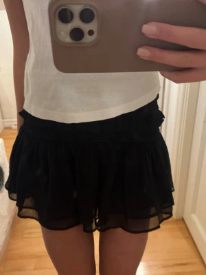 Svart volangskort med resårmidja - Supersöt svart skort med volanger och smockad resårmidja. Luftigt och flowy material som ger en kjol-look men med shortsfunktion. 