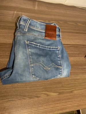 Ljusblå slitna jeans från Replay - Säljer ett par ljusblå jeans från Replay med slitningar och hål på framsidan. Jeansen har klassisk femficksmodell, raka ben och en snygg läderpatch med logga bak i midjan. Perfekta för en avslappnad och trendig stil.