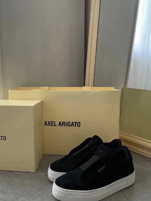 Svarta sneakers från Axel Arigato - Stilrena svarta sneakers från Axel Arigato i mocka med vit tjock sula. Diskret logga på sidan och minimalistisk design utan snörning. Perfekta för dig som gillar en clean och modern look.