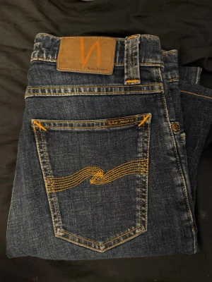 Mörkblå jeans från Nudie Jeans - Säljer ett par mörkblå jeans från Nudie Jeans med klassisk orange kontrastsöm och vågig brodyr på bakfickan. Jeansen har normal passform och är tillverkade i slitstarkt denimtyg. Perfekta för dig som gillar stilrena och tidlösa jeans. Storlek 28/32 slim fit