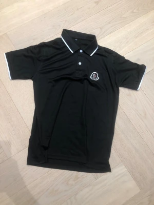 Svart Moncler pikétröja med vit kant - Snygg svart pikétröja från Moncler med vit kant på krage och ärmslut. Klassisk pikéknäppning med två knappar och Moncler-logga broderad på bröstet. Perfekt till jeans eller shorts för en clean och stilren look.