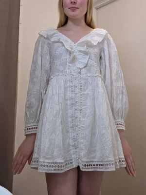 Vit kort studentklänning - Vit kortklänning med volangkrage och spetsdetaljer. Klänningen har långa ärmar, knappar framtill och dekorativa broderade band. Säljer då den tyvärr är lite för kort för mig som är 172cm. Yttermaterialet är 93% bomull och 7%viskos och fodret är 100% bomull.