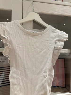 Vit ribbad topp med volangärmar Zara - Superfin vit ribbad topp från Zara med volangdetaljer på ärmarna. Modellen är croppad och har rund halsringning. Perfekt för dig som gillar en söt och trendig stil.