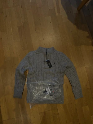  Polo Ralph Lauren quarter zip - Snygg grå kabelstickad tröja från Polo Ralph Lauren med halv dragkedja och ribbade muddar. Tröjan har det klassiska broderade logotypen på bröstet och är tillverkad i mjukt material. Perfekt för en stilren och avslappnad look.