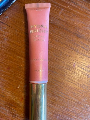 Hydra Bright Cream Blush Peach - Hydra Bright Cream Blush från Revolution Pro i färgen Peach. Krämrouge i tub med skimrande persikoton och guldlock. Perfekt för att ge kinderna en fräsch och naturlig lyster. Enkel att applicera och blanda ut för ett mjukt resultat.