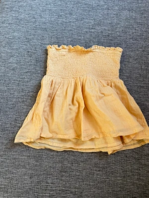 Gul bandeau tubtopp med volang - Supersöt gul bandeau tubtopp med smock upptill och volangig nederdel. Toppen är ärmlös och har en luftig, flowy passform som passar perfekt till sommaren. Tillverkad i ett lätt och mjukt tyg, troligen bomull eller linne.