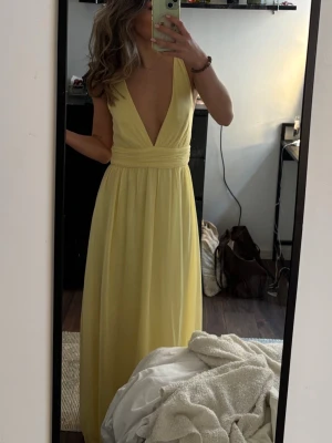 Ljusgul långklänning med v-ringning - Säljer en ljusgul långklänning med djup v-ringning och bred midjedetalj. Klänningen är ärmlös och har en flowy, elegant siluett som passar perfekt till sommarens alla tillfällen. Materialet känns lätt och luftigt.