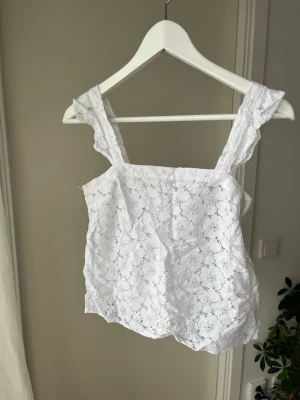 Vit spetslinnetopp med breda axelband - Söt vit linnetopp med breda axelband och vackert spetsmönster. Toppen har en rak skärning och är ärmlös, perfekt för varma dagar. Materialet är lätt och luftigt, vilket ger en fräsch känsla.