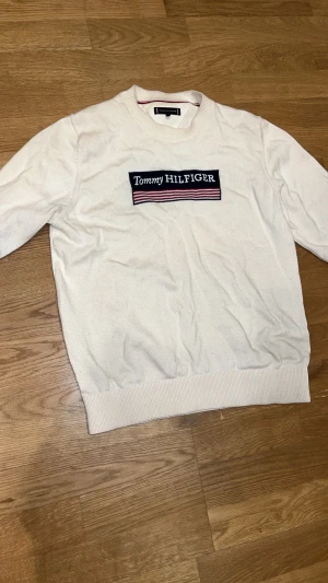 Vit tröja från Tommy Hilfiger - Vit tröja från Tommy Hilfiger med broderad logga på bröstet och ribbade muddar vid hals, ärmslut och nederkant. Klassisk och stilren design med diskreta ränder i kragen. Perfekt för dig som gillar en clean look.