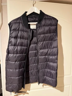 Svart dunväst med ribbad krage - Snygg svart dunväst med quiltad design och ribbad krage. Västen har dragkedja framtill och en stilren look som passar perfekt till lager-på-lager outfits. Materialet är lätt och ger bra värme utan att kännas klumpig.