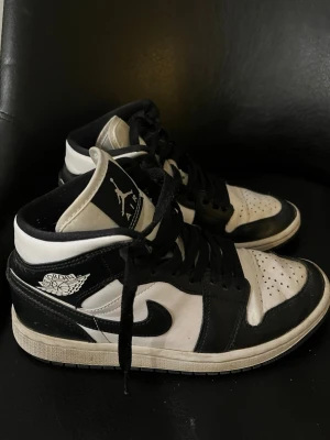Air Jordan 1 Mid svart/vit - Säljer ett par Air Jordan 1 Mid sneakers i svart och vitt läder med klassisk Jordan-logga på sidan och tungan. Skorna har svart swoosh, svarta skosnören och perforerad tåbox för extra ventilation. Perfekt för dig som gillar streetwear och ikonisk design.