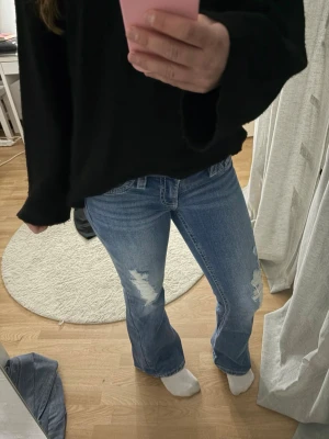 Blå bootcut jeans med slitningar - Säljer ett par ljusblå bootcut jeans med slitna detaljer på låren och klassiska fickor bak med knappstängning. Jätte snygga och aldrig använda! Storlek S från Gina