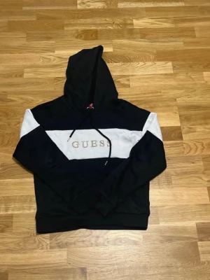 Svart och vit hoodie från Guess - Snygg svart hoodie från Guess med vit panel över bröstet och guldbroderad logga. Klassisk huva med dragsko och mjukt material på insidan. Perfekt för en chill och trendig look.