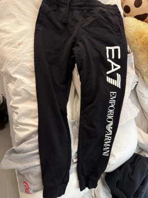 Svarta EA7 Emporio Armani mjukisbyxor - Säljer ett par svarta mjukisbyxor från EA7 Emporio Armani med vit logga längs benet och på bakfickan. Byxorna har resår i midjan och vid bensluten samt snörning i midjan. Perfekta för en avslappnad och trendig look.