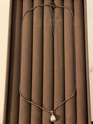 Silverhalsband med pärlhänge - Snyggt och minimalistiskt halsband i silverfärgad metall med ett droppformat vitt pärlhänge. Kedjan är tunn och har justerbar längd med karbinlås och förlängningskedja. Perfekt för dig som gillar stilrena smycken med en elegant touch.