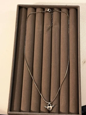Halsband med hjärthängsmycke i silver - Snyggt halsband i silverfärg med ett hjärthängsmycke som har en nyckelhålsdetalj och små vingar på sidorna. Kedjan är tunn och smidig, perfekt för att ge en cool touch till din outfit. Låset är klassiskt och justerbart för bästa passform.