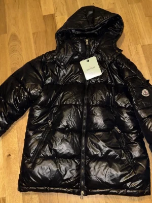 Svart dunjacka från Moncler - Snygg svart dunjacka från Moncler med glansig finish, huva och dragkedja framtill. Jackan har två snedställda fickor med dragkedja och Moncler-logga på ärmen. Perfekt för kalla vinterdagar. Storlekslapp saknas men taggar osv fungerar utmärkt