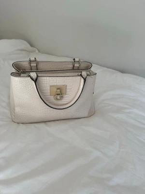 Vit handväska med krokopräglat mönster - Snygg beige handväska med krokopräglat mönster och guldfärgad metallogo framtill. Väskan har två handtag, dragkedja upptill och svarta detaljer längs kanterna. Perfekt för dig som vill ha en stilren och trendig accessoar.