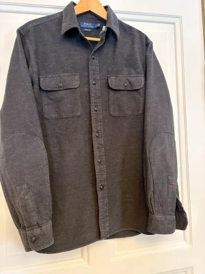 Polo Ralph Lauren overshirt   - Säljer denna riktigt stiliga mörkgrå polo Ralph Lauren overshirt👌 Storlek=M, mycket bra skick: 9,5/10, Inga slitage eller några defekter👏. Vid funderingar är det bara att höra av sig, svarar inom kort👍