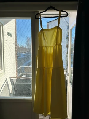 Gul långklänning med smala axelband - Säljer en solgul långklänning med smala axelband och luftig passform. Klänningen har flera volanger och en öppen rygg.