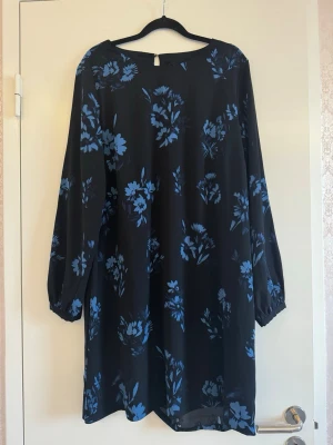 Svart klänning med blå blommor - Svart kortklänning med blått blommönster och långa ärmar. Klänningen har en rak passform och är tillverkad i ett lätt, flödande material som ger en avslappnad look. Perfekt för dig som gillar stilrena men ändå färgglada plagg.