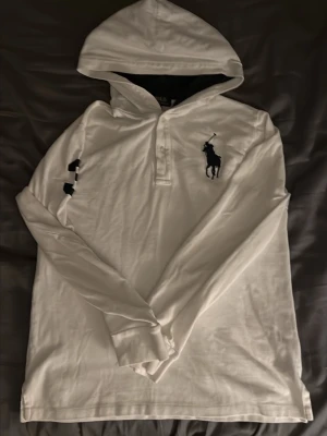 Raphl lauren  - Vit långärmad pikétröja från Ralph Lauren med klassisk krage och knäppning framtill. Stor broderad marinblå logga på bröstet. Perfekt för en clean och stilren look. Riktigt skön och passar fint med en vitt T-shirt under.