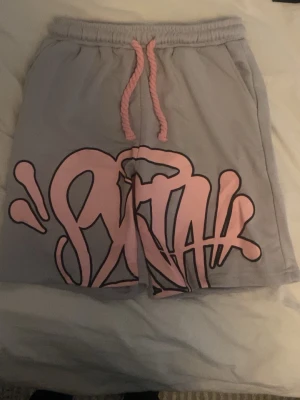 Ljusgrå shorts från syna med tryck - Säljer ett par ljusgrå shorts från syna med stort rosa graffiti-tryck framtill och rosa snörning i midjan. Shortsen har en avslappnad passform och är perfekta för sommardagar. Materialet är mjukt och bekvämt, troligen bomull eller en bomullsblandning.