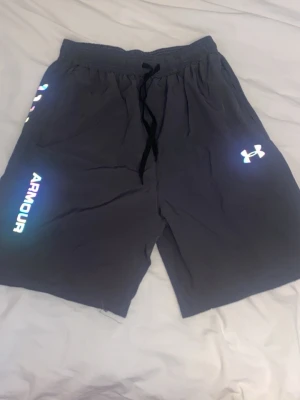 Svarta shorts från Under Armour - Snygga svarta shorts från Under Armour med reflexdetaljer och logga på ena benet. Shortsen har elastisk midja med snörning och är perfekta för träning eller chill. Tillverkade i ett lätt och bekvämt material.