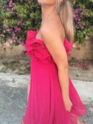  Trendig Axelbandslös rosa volangklänning - Säljer en axelbandslös kortklänning i stark rosa med volangdetaljer upptill. Klänningen har en luftig och flowy passform, perfekt för varma dagar. Materialet känns lätt och svalt, och volangerna ger en extra touch av stil. 
