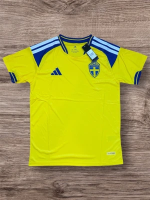 Sverige matchtröja Adidas gul/blå - Gul matchtröja från Adidas med Sveriges landslagsemblem på bröstet och blå detaljer på axlar och ärmslut. Tröjan har korta ärmar, rund hals och är tillverkad i ett lätt, ventilerande funktionsmaterial. Perfekt för fotboll eller träning.
