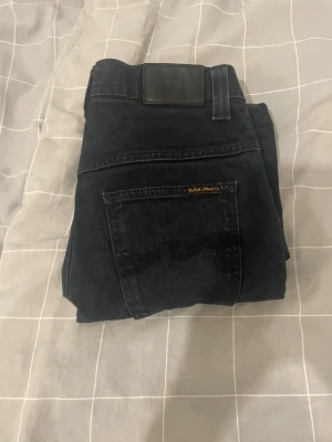 Svarta slim fit jeans från Nudie - Säljer ett par svarta slim fit jeans från Nudie Jeans med klassisk femficksdesign och diskret logga på bakfickan. Jeansen har en smal passform och är tillverkade i mjuk denim med stretch för extra komfort. Kan gå ner i snabbaffär!!
