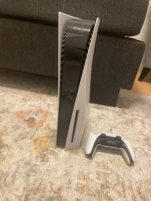 PlayStation 5 med handkontroll - PlayStation 5 konsol från Sony i vit och svart design med futuristiska former. Medföljer en matchande DualSense-handkontroll. Konsolen har en slimmad, stående form och är tillverkad i plast. Kommer med laddare och kablar till den