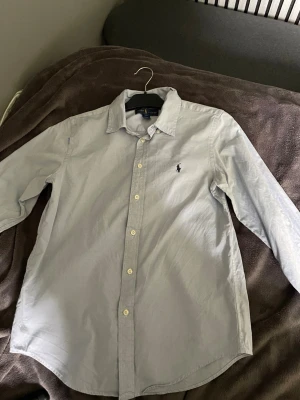 Ljusblå skjorta från Ralph Lauren - Klassisk ljusblå skjorta från Ralph Lauren med broderad logga på bröstet. Skjortan har lång ärm, knappar framtill och en tidlös krage. Perfekt för en clean och stilren look.