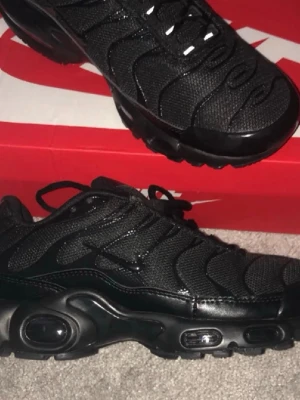 Nike Air Max Plus helsvarta sneakers - Säljer ett par helsvarta Nike Air Max Plus sneakers med mesh- och syntetöverdel, ikoniska vågmönster och synlig Air-dämpning i sulan. Skorna har svarta snören och diskret Swoosh på sidan. Perfekta för dig som gillar streetwear och vill ha en clean look.