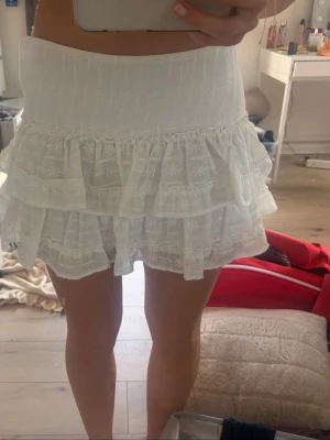 Vit volangkjol hm 36 - Supersöt vit kortkjol med volanger och broderade blommor. Kjolen har en hög midja och är gjord i ett lätt bomullstyg som känns luftigt. Perfekt för varma dagar och ger en romantisk vibe till din outfit. 