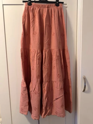 Lång rosa kjol med volanger - Säljer en lång, luftig kjol i en mjuk rosa nyans med volangdetaljer. Kjolen har ett subtilt broderat mönster och resår i midjan för skön passform. Perfekt för dig som gillar en avslappnad men ändå söt stil. HOMEMADE!