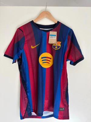 Barcelona matchtröja Nike blå/röd - Snygg FC Barcelona matchtröja från Nike med klassiska blå och röda vertikala ränder. Tröjan har klubbmärke, Nike-logga och Spotify som sponsor framtill samt 'BARCA FOUNDATION' på ryggen. Tillverkad i lätt och ventilerande polyester, perfekt för fotboll eller träning.