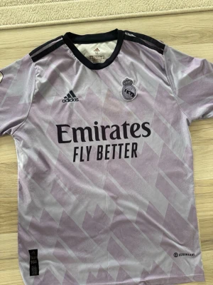 Lila Real Madrid tröja Adidas  - Snygg lila Real Madrid matchtröja från Adidas med svart krage och detaljer. Tröjan har klubbmärke, Adidas-logga och Emirates Fly Better-tryck på bröstet. Tillverkad i lätt och ventilerande AEROREADY-material, perfekt för fotbollsträning eller match.