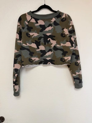 Camo crop hoodie i grönt och rosa - Säljer en croppad hoodie med camouflagemönster i grönt, svart, grått och rosa. Tröjan har långa ärmar, rund halsringning och rå kant nertill för en edgy look. Perfekt för dig som gillar streetwear och vill sticka ut. ( tänk på jag har hund i hemmet) 