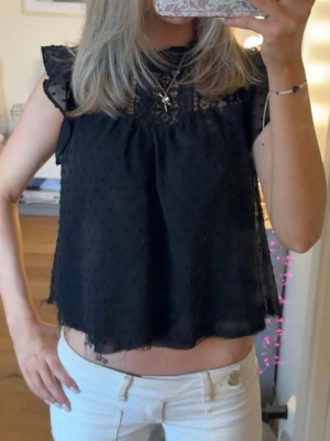 Svart blus med spetsdetaljer - Säljer en svart blus med spets upptill och volangdetaljer vid axlarna. Blusen är ärmlös och har ett lätt transparent tyg med diskret prickigt mönster. Tyvärr är knappen där bak i nacken som man knäpper av men det går att sy dit en💞