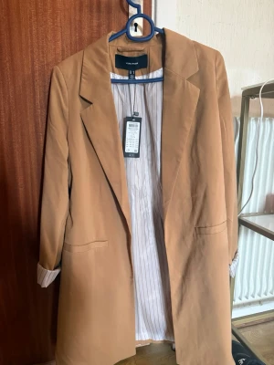 Beige kappa från Vero Moda - Stilren beige kappa från Vero Moda med klassisk krage och raka fickor. Insidan har ett vitt foder med tunna bruna ränder och uppvikta ärmar visar fodret för en snygg detalj. Perfekt för dig som gillar minimalistisk och trendig stil.