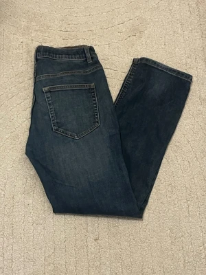Mörkblå raka jeans - Säljer ett par mörkblå jeans med rak passform och klassiska bakfickor. Jeansen har en enkel design utan slitningar eller dekorationer och är tillverkade i denim. Perfekta för dig som gillar en clean och tidlös look.