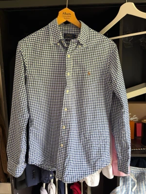 Blårutig skjorta från Polo Ralph Lauren - Klassisk blårutig skjorta från Polo Ralph Lauren i slim fit-modell. Skjortan har långa ärmar, knappar framtill och den ikoniska broderade loggan på bröstet. Tillverkad i mjuk bomull och passar perfekt till både jeans och chinos.