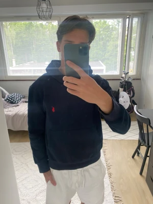 Mörkblå hoodie från Polo Ralph Lauren - Snygg mörkblå hoodie från Polo Ralph Lauren med klassisk röd logga broderad på bröstet. Hoodien har huva och en stor magficka framtill. Tillverkad i mjukt bomullsmaterial som känns skönt mot huden. Perfekt för en avslappnad och stilren look.