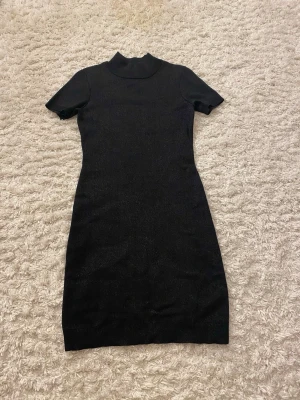 Svart glittrig  klänning - Säljer en svart  klänning med glittrigt tyg och korta ärmar. Klänningen har en hög krage och är figurnära i modellen. Perfekt för dig som gillar en enkel men ändå festlig look.