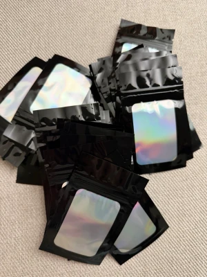 50st Svarta zip-påsar med holografiskt fönster, 10x6 - 50 stycken  små zip-påsar i svart plast med ett genomskinligt holografiskt fönster på framsidan. Perfekta för förvaring av småsaker, smycken eller andra prylar. Påsarna är återförslutningsbara och har en modern, glansig look.  Storlek: 10x6