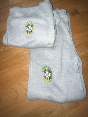 Brazilien tracksuit. - Säljer en grå hoodie med huva och magficka, samt matchande byxor. Båda plaggen har Brasilien-logga med stjärnor på bröstet och benet. Perfekt för dig som gillar sportig stil och vill representera Brasilien. Mjuk och bekväm bomullsblandning.
