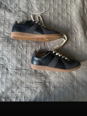 Maison margiela gats - Stilrena svarta/gråa sneakers från Maison margiela med detaljer i mocka och släta partier i läder. Skorna har vita snören och en klassisk gummisula i brunt. Perfekt för dig som gillar minimalistisk och tidlös design.