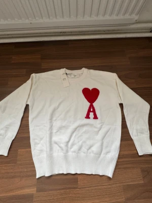 Ami - Knitwear - Vit stickad tröja från Ami Paris med röd hjärtlogga och bokstaven A på bröstet. Tröjan har rund halsringning, ribbade muddar och är tillverkad i mjukt material. Perfekt för dig som gillar stilrena plagg med en twist. Inga defekter